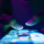 Close-up of hands scrolling on a smartphone screen in a dark environment with blue and purple lighting.
