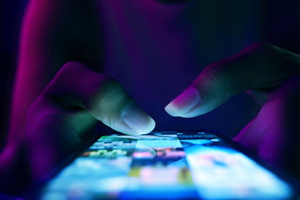 Close-up of hands scrolling on a smartphone screen in a dark environment with blue and purple lighting.