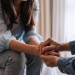 Two people sitting closely, with one person holding the other's hands in a gesture of support and compassion.