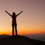 A person standing on a mountaintop at sunset with arms raised triumphantly, symbolizing resilience, freedom, and personal growth.
