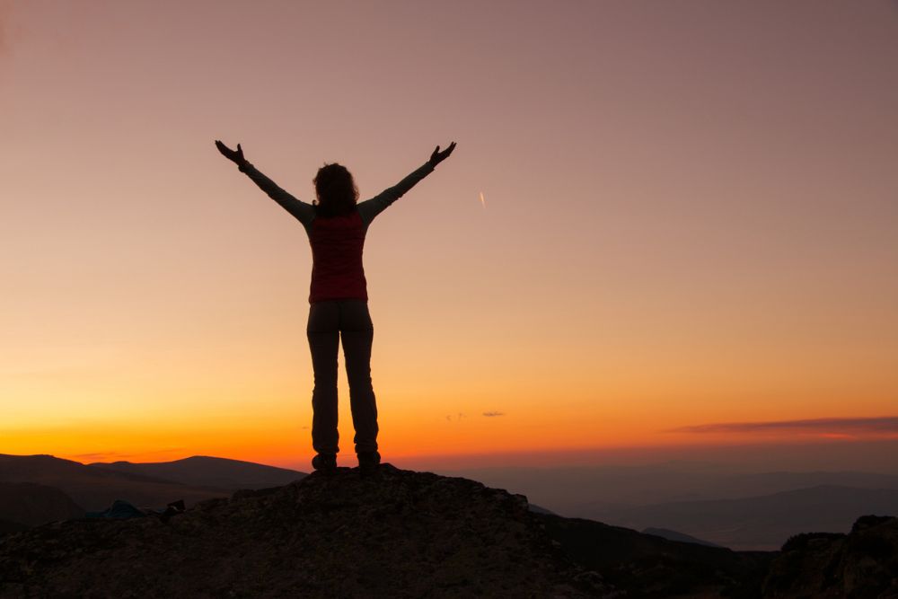 A person standing on a mountaintop at sunset with arms raised triumphantly, symbolizing resilience, freedom, and personal growth.