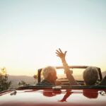 Two older adults in a red convertible enjoying a scenic drive at sunset, with one person raising their arms joyfully into the air, symbolizing freedom and happiness in retirement.
