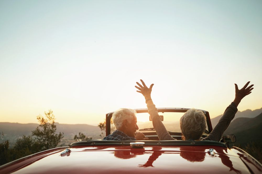 Two older adults in a red convertible enjoying a scenic drive at sunset, with one person raising their arms joyfully into the air, symbolizing freedom and happiness in retirement.