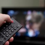 A hand holds a TV remote, pressing the power button to turn off the television. The screen shows a blurred image of people in the background.