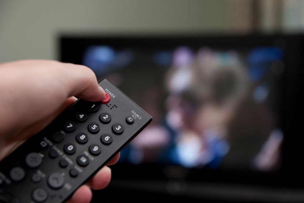 A hand holds a TV remote, pressing the power button to turn off the television. The screen shows a blurred image of people in the background.