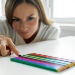 A woman carefully arranges colored pencils in a neat row on a white table, focusing intently, symbolizing perfectionism or obsessive behavior.