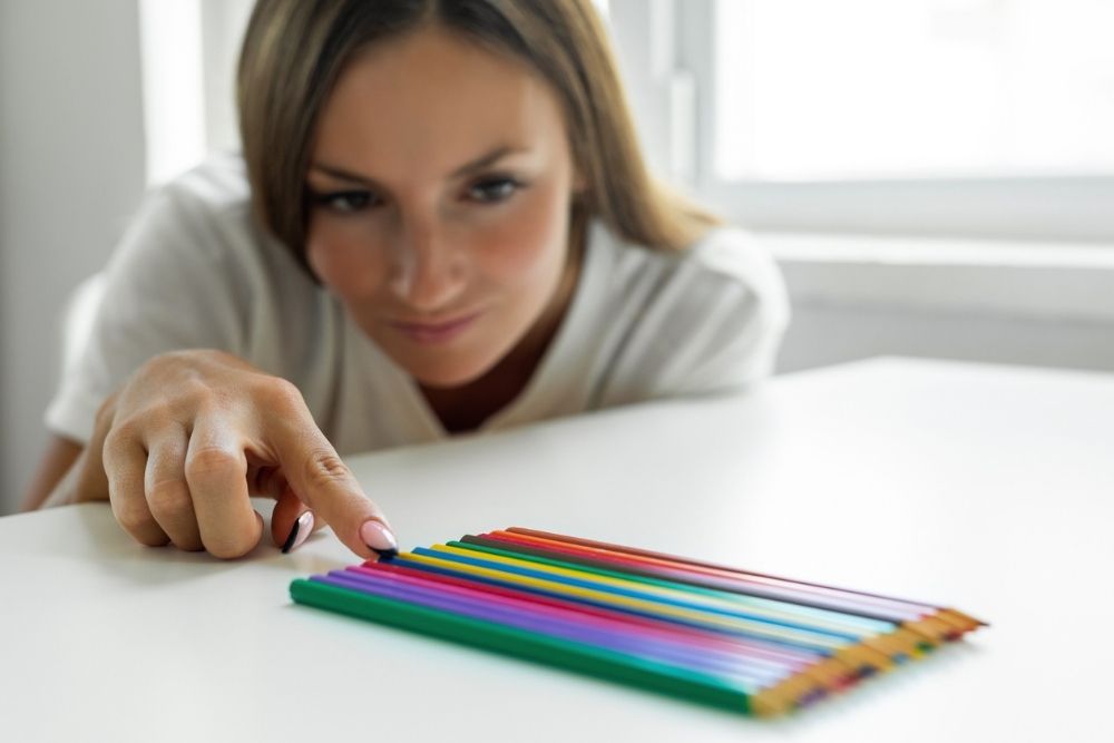 A woman carefully arranges colored pencils in a neat row on a white table, focusing intently, symbolizing perfectionism or obsessive behavior.