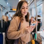 Young woman standing on public transit while looking at her phone, symbolizing daily social media use and digital connection.