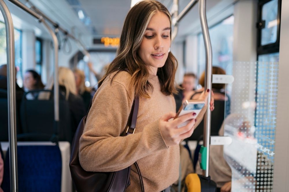 Young woman standing on public transit while looking at her phone, symbolizing daily social media use and digital connection.