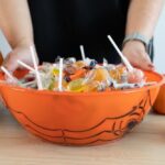 A person holds an orange bowl filled with assorted Halloween candy, surrounded by decorative jack-o’-lanterns.
