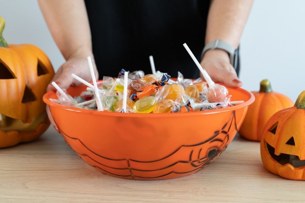 A person holds an orange bowl filled with assorted Halloween candy, surrounded by decorative jack-o’-lanterns.