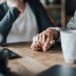 Close-up of two people sitting at a table, gently holding hands in a comforting gesture, with a coffee mug and phone nearby.