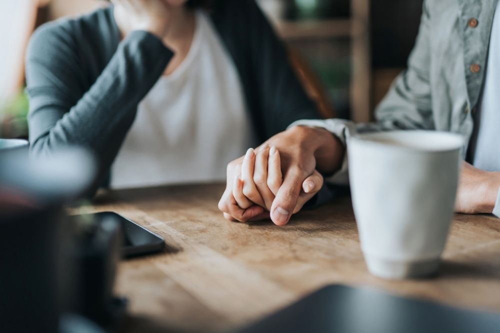 Close-up of two people sitting at a table, gently holding hands in a comforting gesture, with a coffee mug and phone nearby.