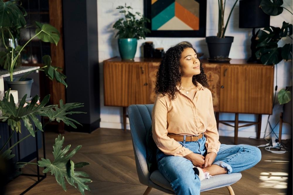 A woman sitting cross-legged in a chair with her eyes closed, taking a deep breath and practicing mindfulness in a peaceful, plant-filled room.
