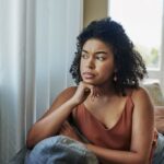 A woman sits on a couch looking out the window with a thoughtful, concerned expression, symbolizing negative self-talk or self-doubt.