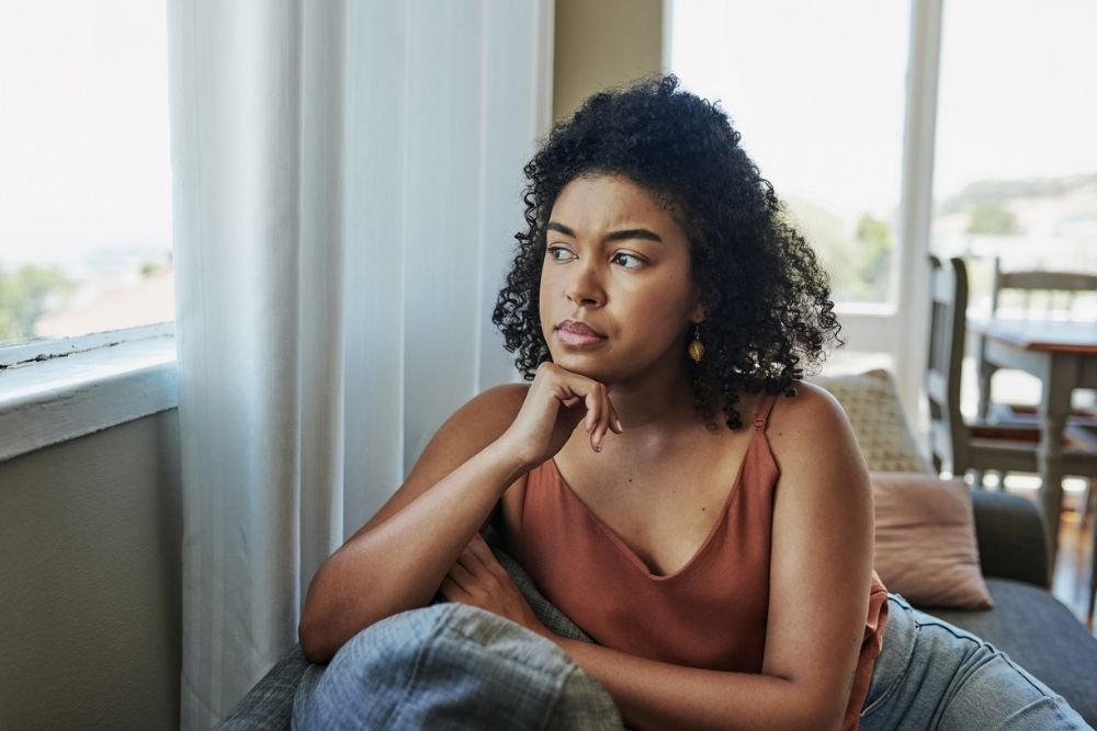 A woman sits on a couch looking out the window with a thoughtful, concerned expression, symbolizing negative self-talk or self-doubt.