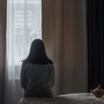 A person sitting on a bed facing a window with curtains drawn, appearing reflective or sad, while a cat rests beside them.