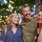 A group of middle-aged friends stand close together outdoors, smiling and laughing during a casual social gathering.