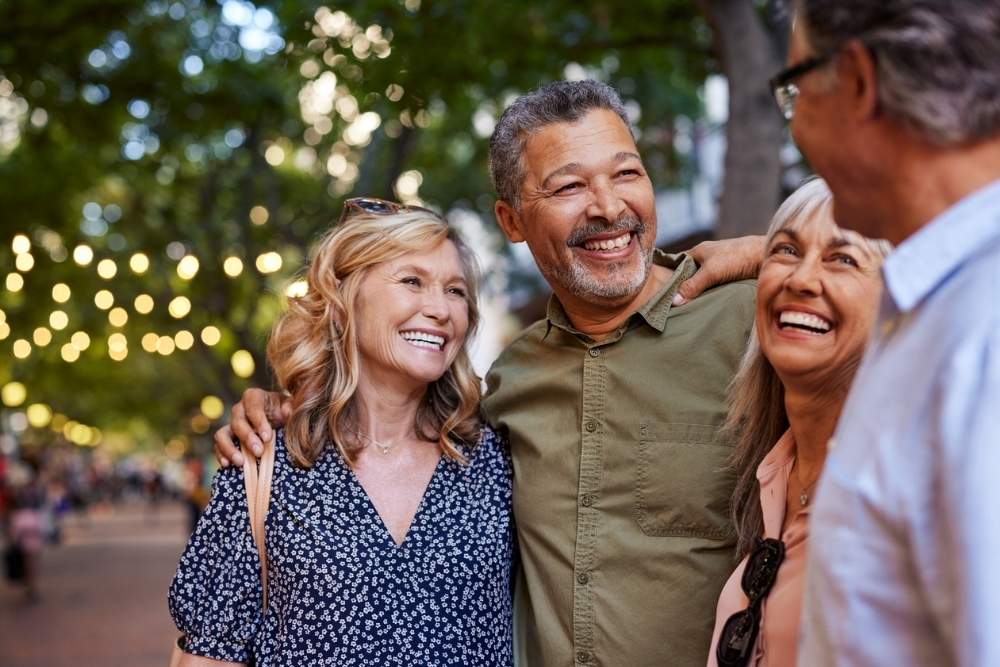 A group of middle-aged friends stand close together outdoors, smiling and laughing during a casual social gathering.
