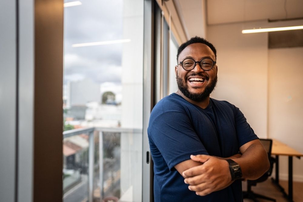 Person smiling confidently with arms crossed near a window, appearing calm and positive in a modern indoor setting.