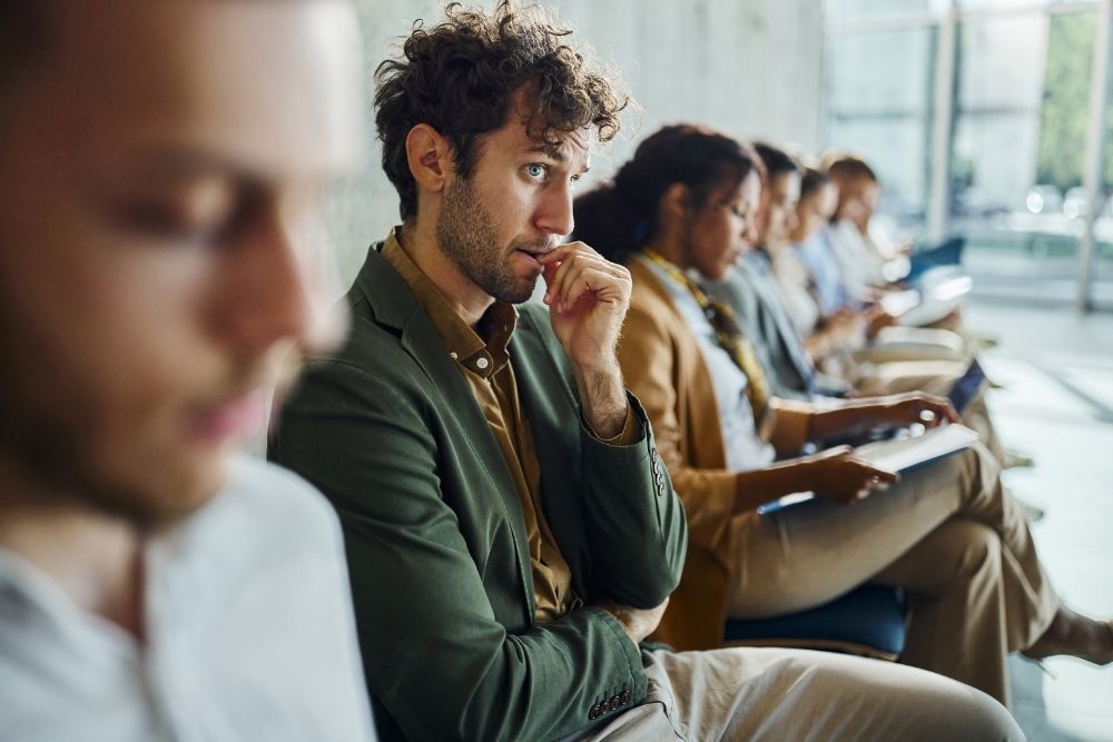 A group of people seated in a waiting area, with one person with high-functioning anxiety is in focus biting their fingernail in apparent contemplation.