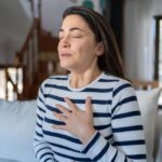 Palms - What Makes Panic Attacks Happen - Edited. A woman sits on a coach holding her palm to her chest. She appears to be taking a deep breath.