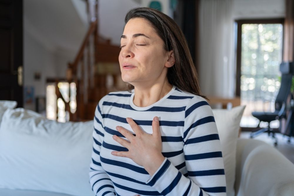 Palms - What Makes Panic Attacks Happen - Edited. A woman sits on a coach holding her palm to her chest. She appears to be taking a deep breath.